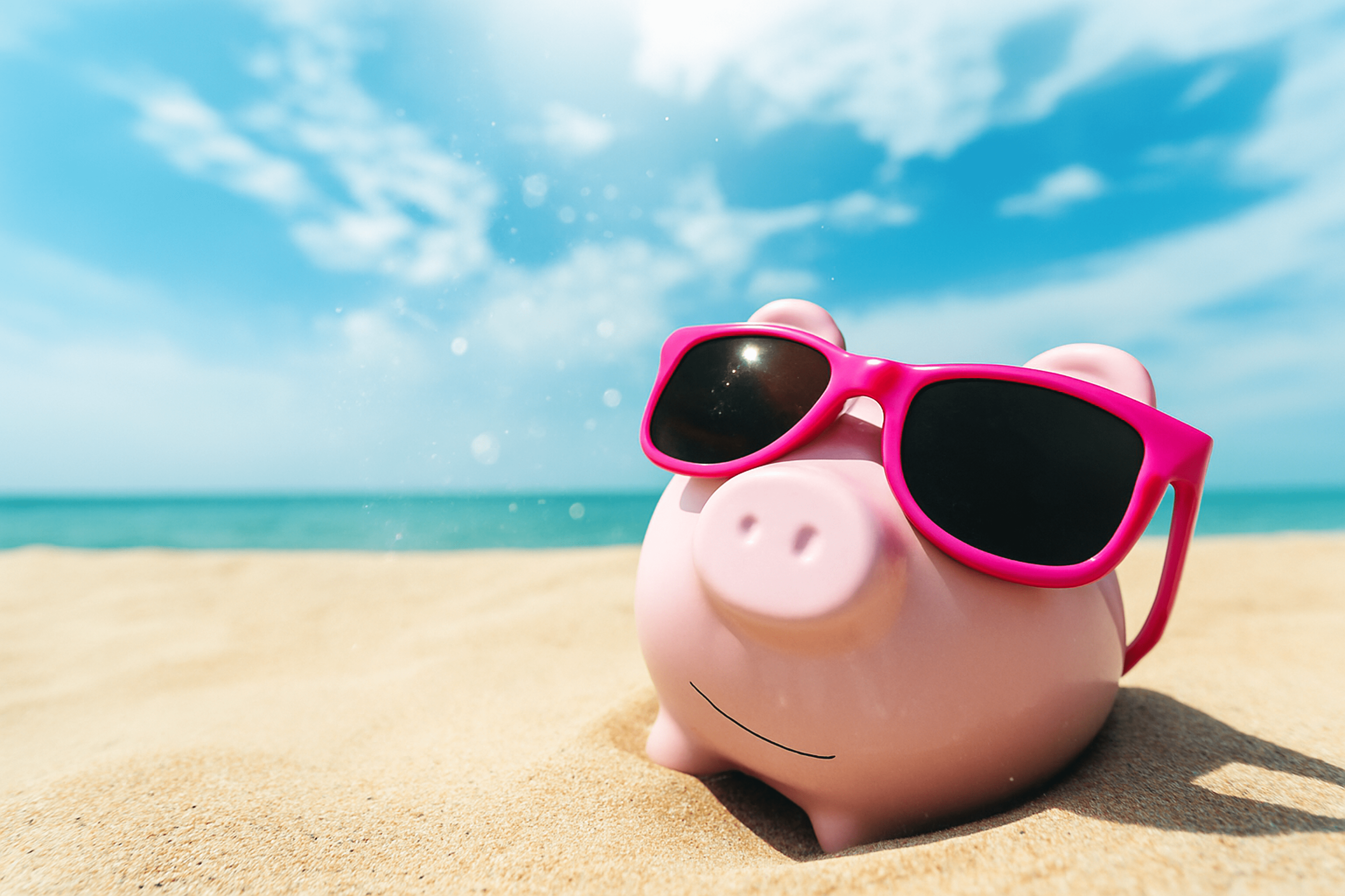 A pink piggy bank wearing pink sunglasses sits on the sand at the beach under a blue sky.